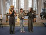 Kind spreekt in microfoon in de kerk. Twee vrouwen staan ernaast, één houdt de microfoon vast.