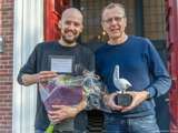 Twee glimlachende mannen houden de Pelikaan-prijs en een bos bloemen vast.